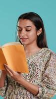 Vertical Cheerful woman smiling while enjoying reading activity, holding book, isolated over studio background. Radiant person pleased with entertaining novel, enjoying leisure time video