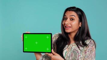 Happy woman presenting green screen tablet, isolated over studio background. Joyous indian person holding copy space chroma key device used for advertising brands, camera B video