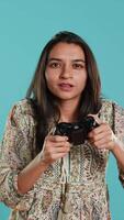 Vertical Cheerful indian gamer showing thumbs up sign gesturing while holding controller after winning videogame. Joyful woman enjoying games, doing approving hand gesture, studio background, camera A video