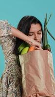 vertikal kvinna med papper väska i händer fylld med grönsaker kontroll morot för föroreningar. medveten levande kund med köpt specerier till vara Begagnade som matlagning Ingredienser, studio bakgrund, kamera en video