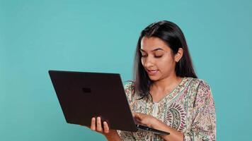 mujer composición correo electrónico, cuidadosamente mecanografía en ordenador portátil teclado, luchando a pensar de qué a escribir. indio persona enviando correo en línea, lluvia de ideas mensaje idea, estudio fondo, cámara si video