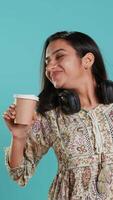 Vertical Joyful indian woman stretching and sipping fresh coffee from disposable paper cup after waking up. Radiant lively person drinking hot beverage early in the morning, studio background, camera A video