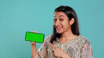 Smiling woman showing isolated screen mobile phone, doing promotion, studio background. Upbeat indian person holding copy space mockup cellphone used for advertising brands, camera B video