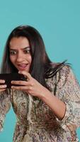 Vertical Happy woman playing videogames on smartphone, celebrating after defeating foes, doing clenching hand gesture. Player swiping phone screen, thrilled about winning game, studio background, camera A video