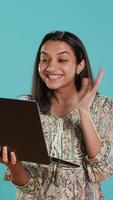 Vertical Upbeat woman saluting coworkers, having friendly conversations during teleconference meeting using laptop, studio background. Person discussing during online videocall on notebook device, camera A video