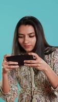 verticale contento indiano donna intrattenuto di videogiochi su smartphone, godendo tempo libero volta. gamer godendo gioco su Telefono, avendo divertimento sconfiggere nemici, isolato al di sopra di studio sfondo, telecamera un' video