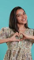 Vertical Portrait of smiling loving woman doing heart symbol shape gesture with hands, showing kindness. Happy affectionate girlfriend doing tender love gesturing for boyfriend, studio background, camera A video