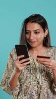 Vertical Indian woman excited to purchasing stuff, adding payment method on mobile phone website, isolated over studio background. Ecstatic client does internet shopping, typing debit card info, camera A video