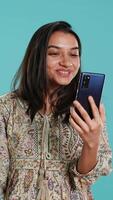 Vertical Smiling woman having friendly conversations during teleconference meeting using smartphone, studio background. Indian person having fun catching up with mates during online videocall, camera A video