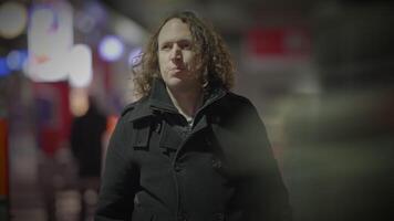 un hombre con largo Rizado pelo es caminando abajo un calle a noche video