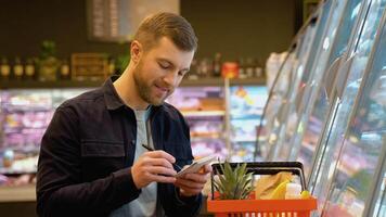 Young man with full shopping basket reading shopping list. A man makes notes in the shopping list video
