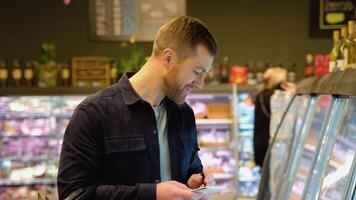 A man makes notes in the shopping list. Young man with trolley full of products reading shopping list video