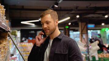 Portrait of confused man doing grocery shopping in supermarket. calling his wife asking what healthy snack of nuts and dried fruit to choose video