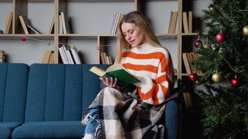 Attractive young lady with blonde hair reading interesting book while sitting on blue couch near beautiful christmas tree. Cozy atmosphere video