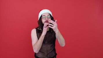 Young sexy gay man in a Santa hat paints lips isolated on red background. People lifestyle fashion lgbtq concept video