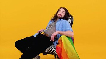 Gay man dressed in mesh t-shirt sits in a chair with a multicolored flag. Concept diversity, transsexual, and freedom video