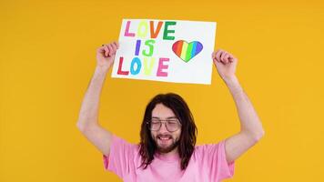 ung aktivist leende Lycklig roligt Gay man i rosa t-shirt innehav en protest tecken under en HBTQ stolthet parad isolerat på gul bakgrund studio. människor HBTQ livsstil begrepp. kärlek är kärlek video