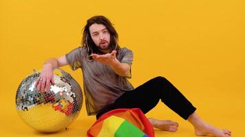Gay man dressed in mesh t-shirt sits on yellow background with a multicolored flag and silver ball. Concept diversity, transsexual, and freedom video