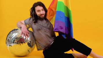 Gay man dressed in mesh t-shirt sits on yellow background with a multicolored flag and silver ball. Concept diversity, transsexual, and freedom video