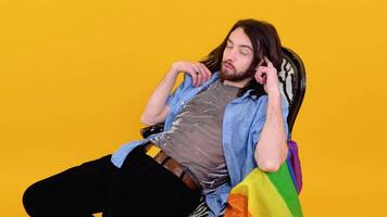 Gay man dressed in mesh t-shirt sits in a chair with a multicolored flag. Concept diversity, transsexual, and freedom video