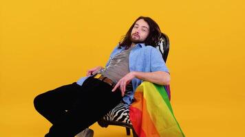 jung kaukasisch Fröhlich Mann 20er Jahre mit Regenbogen gestreift Flagge sitzt im ein Stuhl isoliert auf Gelb Hintergrund. Menschen Lebensstil Mode lgbtq Konzept video