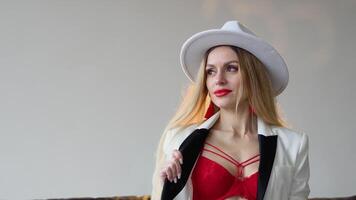 en kvinna med färgade hår och smink i en hatt, vit jacka och röd behå poser i de studio. mode skytte begrepp video