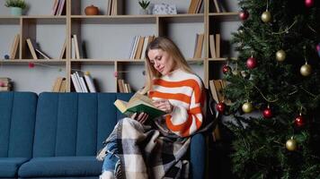 Attractive young lady with blonde hair reading interesting book while sitting on blue couch near beautiful christmas tree. Cozy atmosphere video