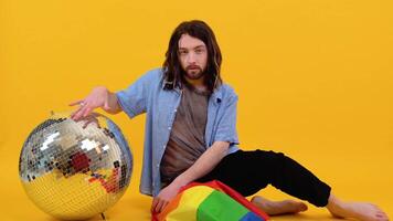 Gay man dressed in mesh t-shirt sits on yellow background with a multicolored flag and silver ball. Concept diversity, transsexual, and freedom video