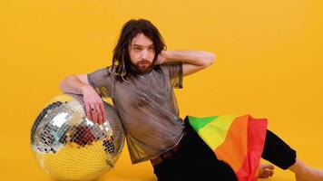 Young caucasian gay man 20s with rainbow and silver ball striped flag sits on yellow background. People lifestyle fashion lgbtq concept video