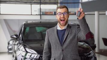 positivo masculino sonriente para cámara y demostrando llaves mientras en pie cerca nuevo vehículo en sala de exposición video