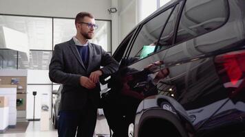 A stylish man in glasses and suit closes the door of the new electric car in car dealership. A man examine vehicle before making purchase. Buy car video