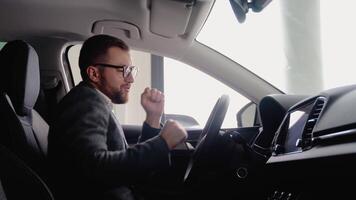 Happy man sits in new car in shop dealership and celebrate purchase of new vehicle. Buy car video