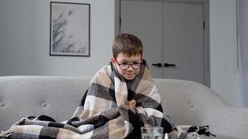 A little boy with flu, fever and headache sitting wrapped in a plaid on the couch. Period of seasonal diseases. Concept of viral diseases video