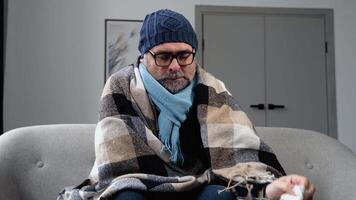 doente congeladas Senior homem dentro uma chapéu sentar em sofá, vestem chapéu detém lenço de papel sopro escorrendo nariz obteve febre, pegar frio, amontoar do remédios perto video