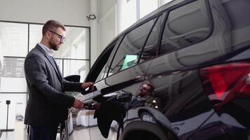 A stylish man in glasses and suit closes the door of the new car in car dealership. A man examine vehicle before making purchase. Buy car video