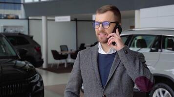 Caucasian man in glasses and suit speaks by phone in car dealership video