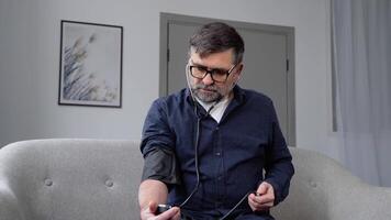 Bearded senior man using modern tonometer at home. A man measuring his blood pressure, making self checkup. Heart problems, heart attack prevention video