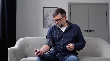 Senior Mann mit modern Tonometer beim heim. ein Mann Messung seine Blut Druck und sieht aus beim das Tonometer im Schreck video