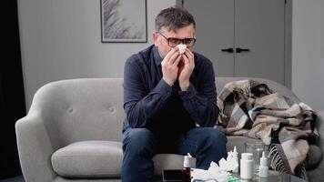 Sick frozen senior man sit on sofa, wear hat holds tissue blowing runny nose got fever, catch cold, heap of meds nearby. Seasonal influenza symptoms, flu, unheated flat concept video