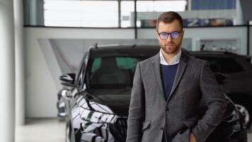 porträtt av en ung man i glasögon och kostym nära en ny bil i en bil återförsäljare video