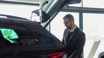 een Mens sluit de romp van een nieuw auto in de auto dealerschap. zakenman onderzoeken luxe voertuig voordat maken aankoop video