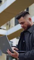 Businessman with a laptop outdoors. Handsome man in warm coat standing in the city street and working on a laptop. Business and work concept. Vertical video