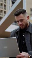 Young man with a laptop on fresh air. Handsome young man in a coat sitting in the street and working on a modern device in the city. Distance work. Vertical video