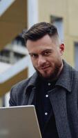 Portrait of a positive man with a laptop. Handsome bearded businessman working on a laptop in the street on the modern building background. Freelance job. Vertical video