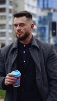 Young man drinking coffee in the street. Handsome bearded man in elegant warm coat walking in the city with a drink in plastic cup. Vertical video