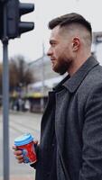 stilig man på urban bakgrund. profil av en ung affärsman med plast kopp stående nära de väg och ser på sidor i de stad. vertikal video