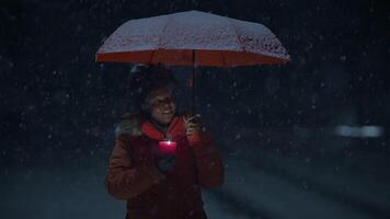 Jeune africain femme permanent sur rue à nuit pendant neige tomber avec parapluie video