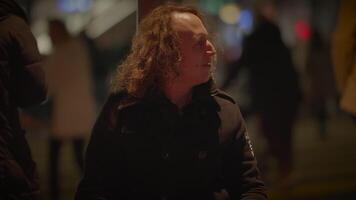 A man with long curly hair is seen in various locations, including in front of a sign, a window, a logo, and in a dark room having a conversation. The also shows him sitting on a bus at night video