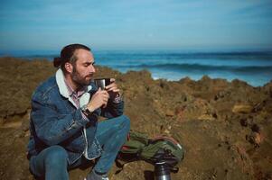 Brutal bearded man have rest, enjoys his coffee break outdoor, sitting with his eyes closed on the rocky cliff by ocean photo