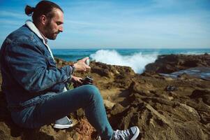 Bearded male tourist relaxing on the rocky beach, enjoying his coffee break outdoor. Active tourism and camping concept photo